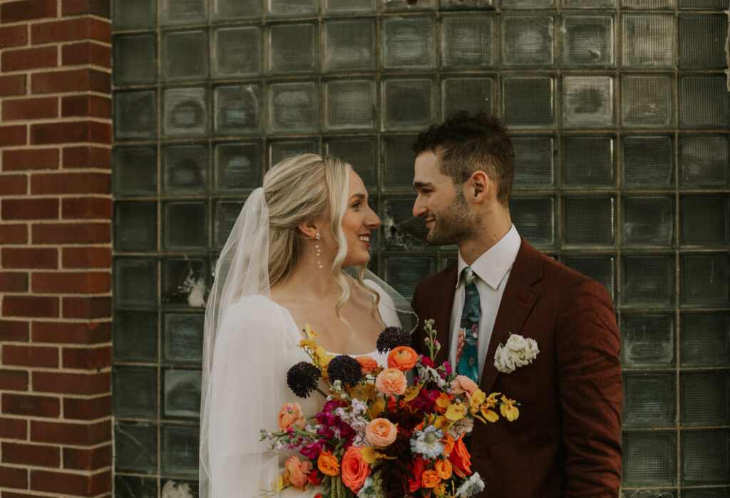 upscale bride and groom portraits outside of historical wedding venue with square glass brick wall and colorful floral bouquet