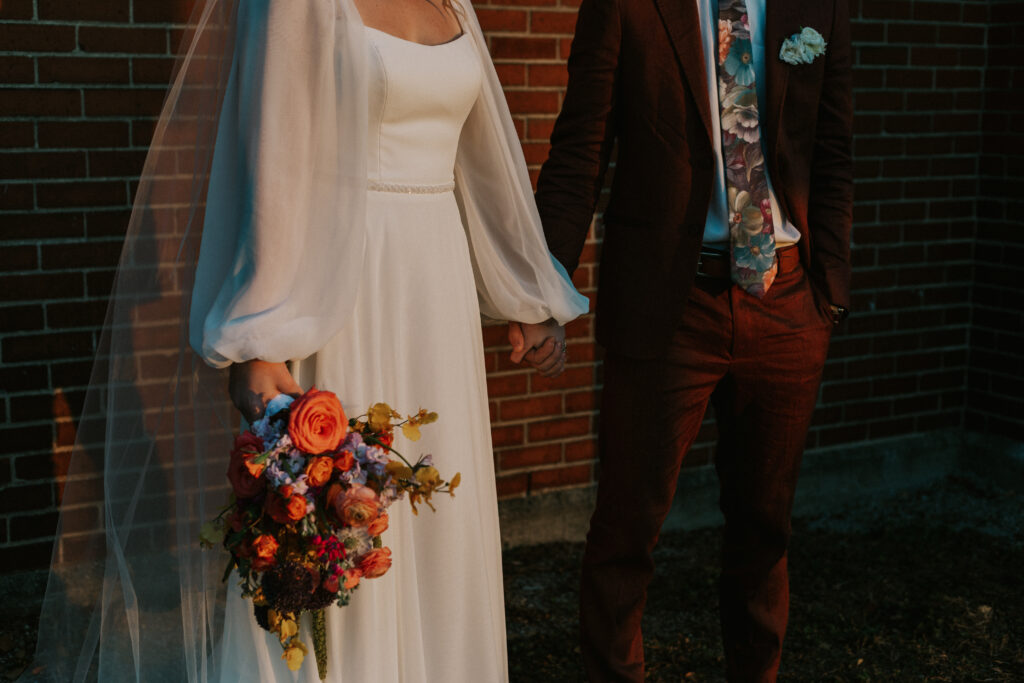 upscale bride and groom portraits outside of historical wedding venue with square glass brick wall and colorful floral bouquet during golden hour