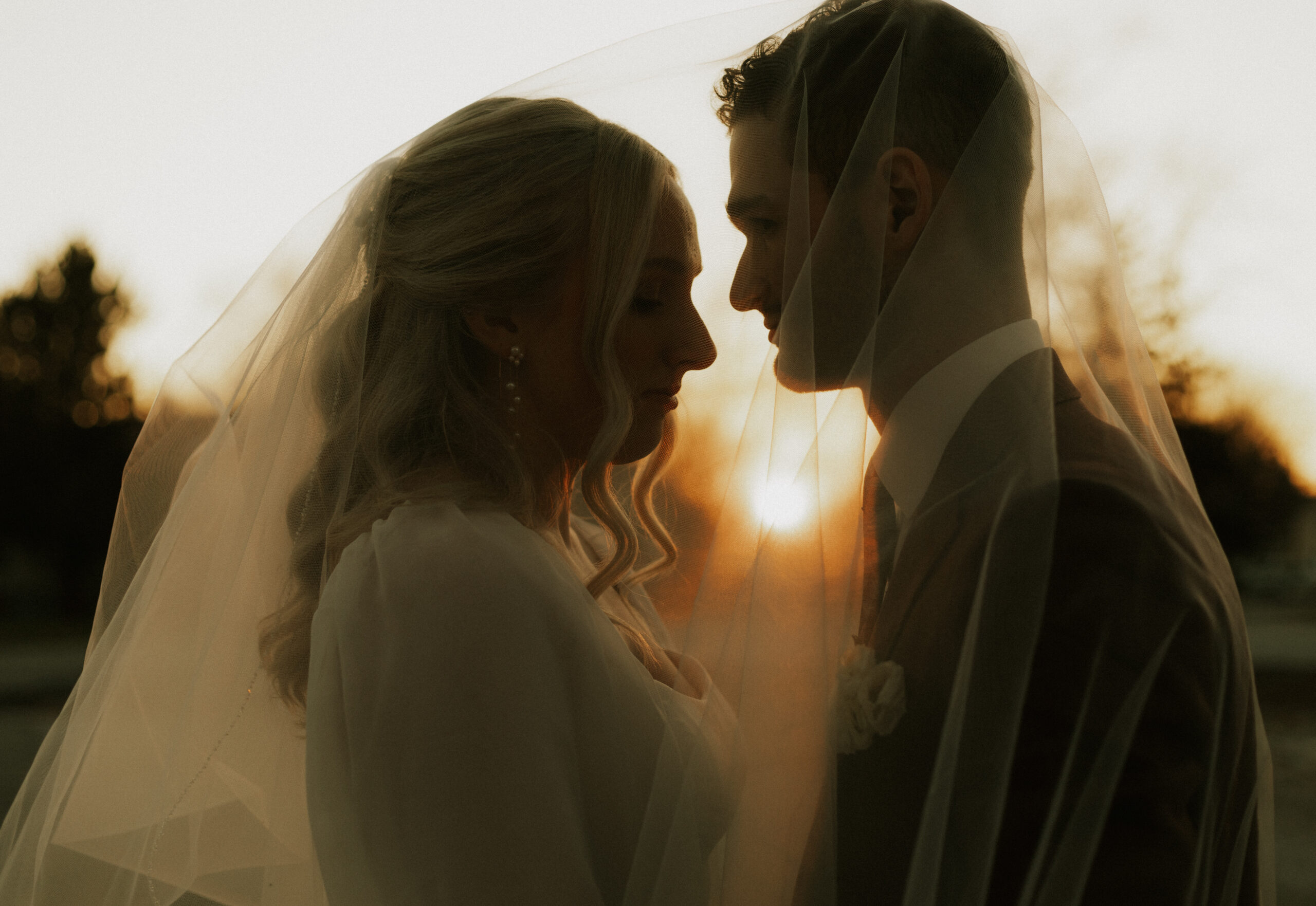 wedding bride and groom underneath veil during golden hour, with sun shining through veil about to kiss