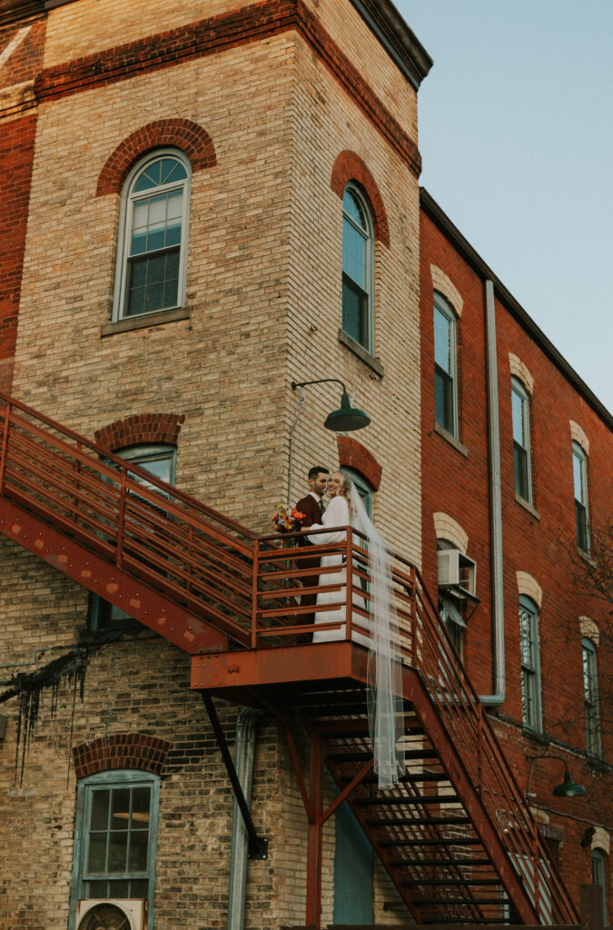 upscale bride and groom portraits outside of historical wedding venue during golden hour on rust colored stairs with veil blowing in wind