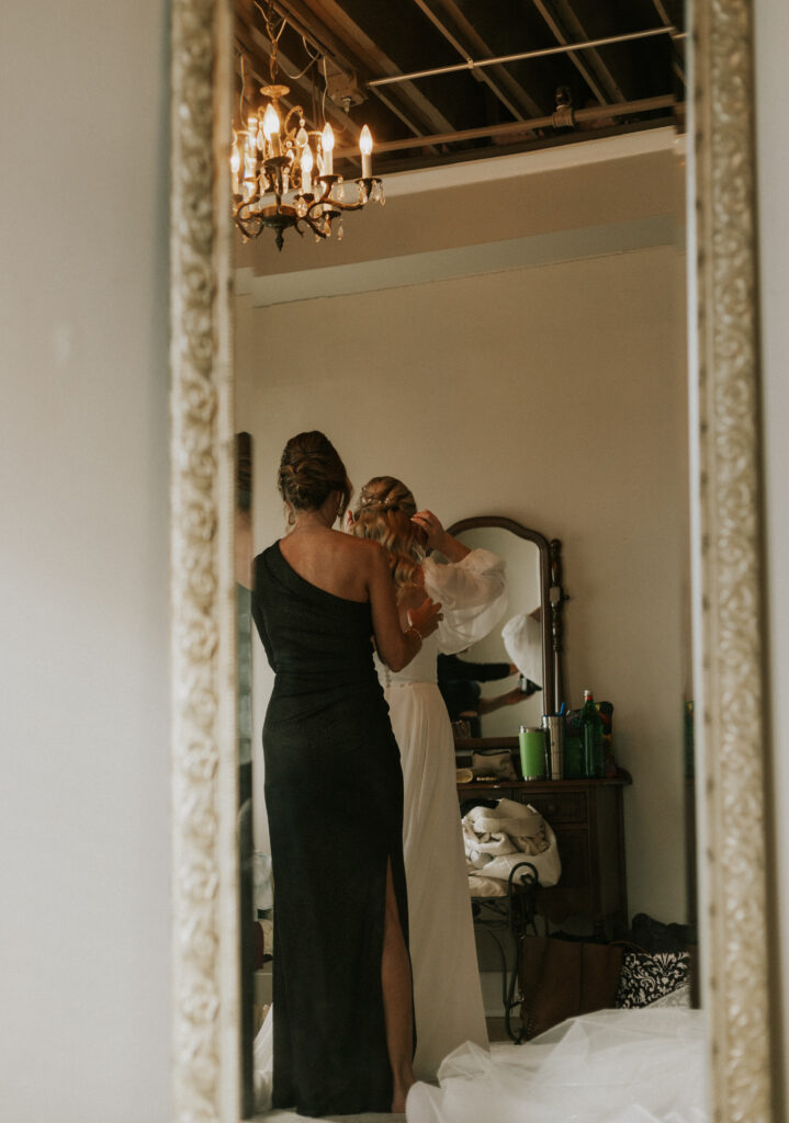 bride getting ready in bridal suite at Bread & Chocolate wedding venue, mom zipping up dress in the mirror reflection
