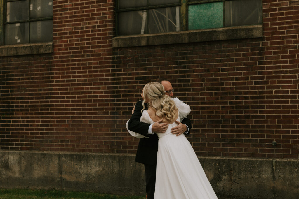 first look with bride and father of the bride in a field with a historical brick building with stained glass, father of bride sees bride for the first time and hug