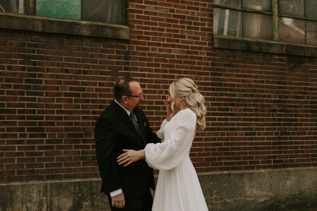 first look with bride and father of the bride in a field with a historical brick building with stained glass, father of bride sees bride for the first time and hug and shed tears