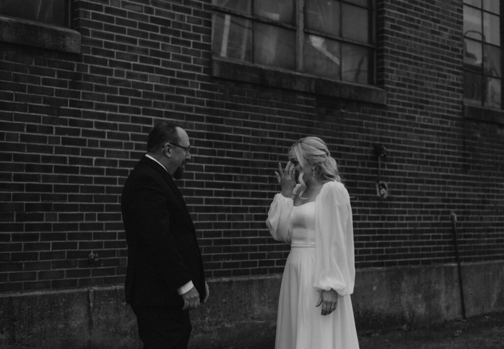 first look with bride and father of the bride in a field with a historical brick building with stained glass, father of bride sees bride for the first time and hug and shed tears together