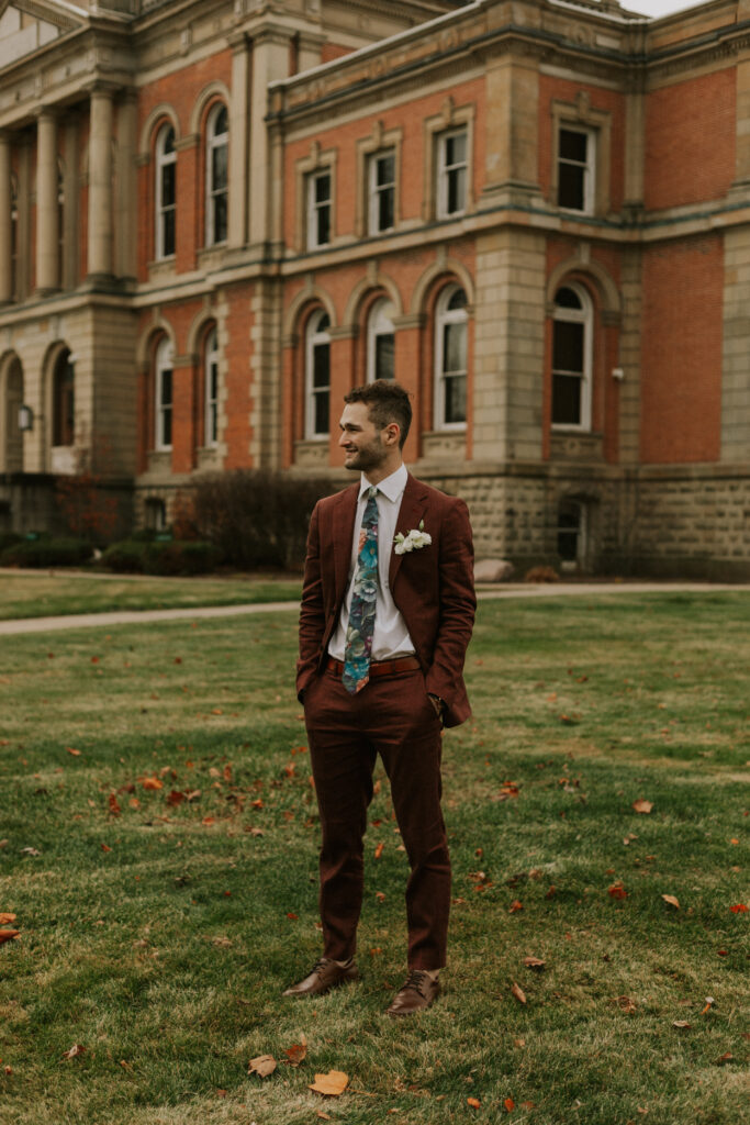 groom portraits at Goshen, Indiana courthouse downtown Goshen on steps with historical building in background with a maroon suit, floral tie, and white floral boutonniere 