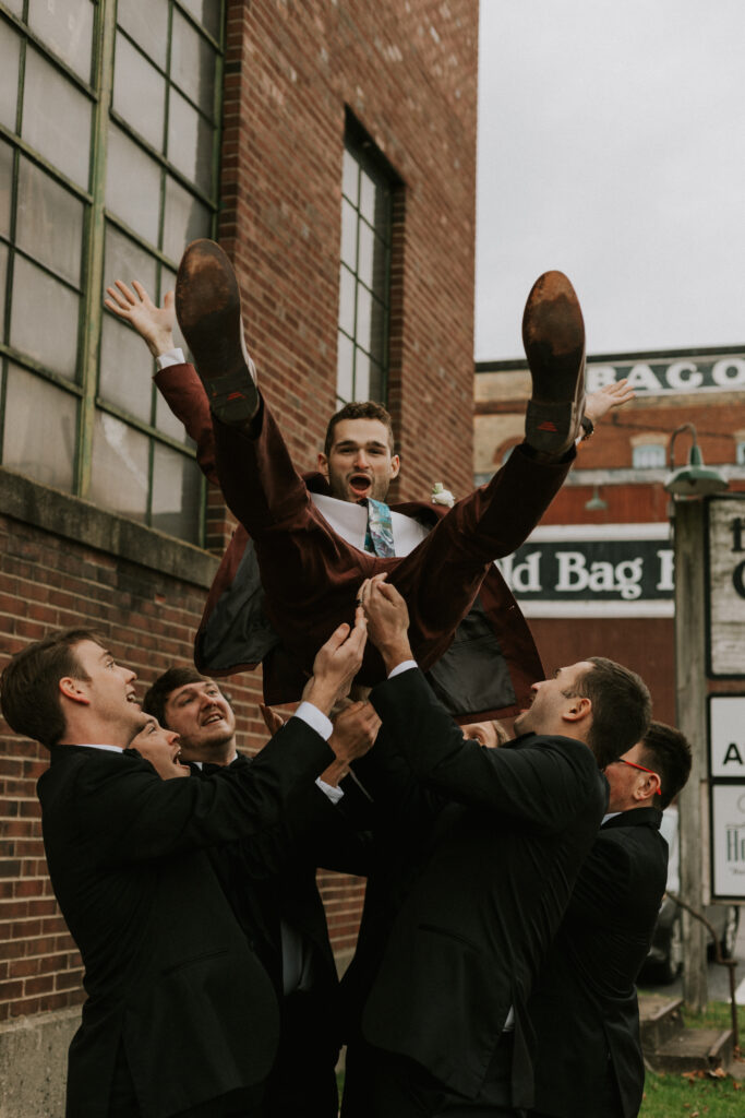 Groomsmen party pictures in front of the historical brick building in downtown Goshen Indiana at Bread & Chocolate, tossing groom in the air pictures