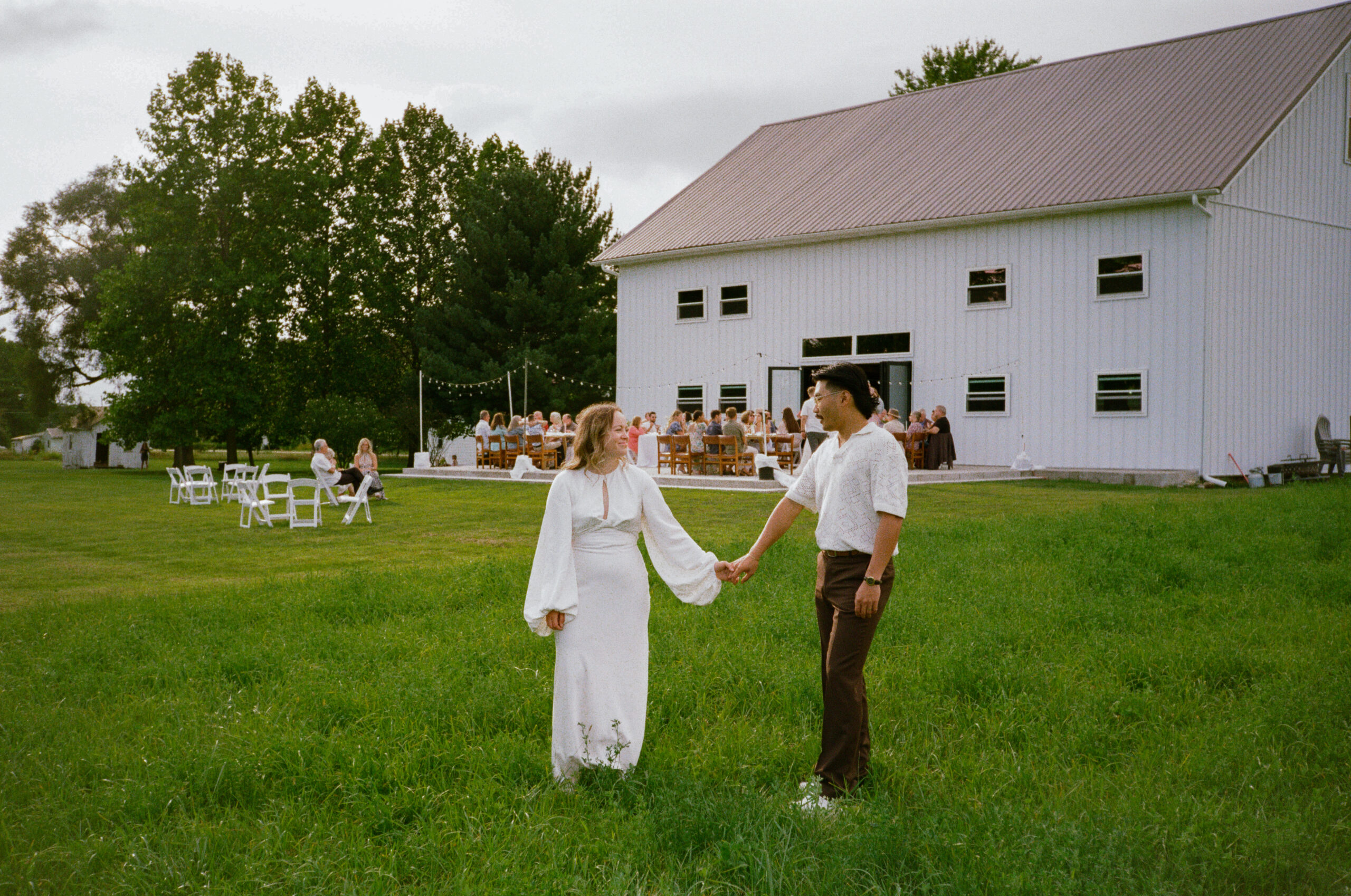 Wedding couple standing in front of outdoor reception space in winona lake, indiana holding hands, guests are in the background on film.