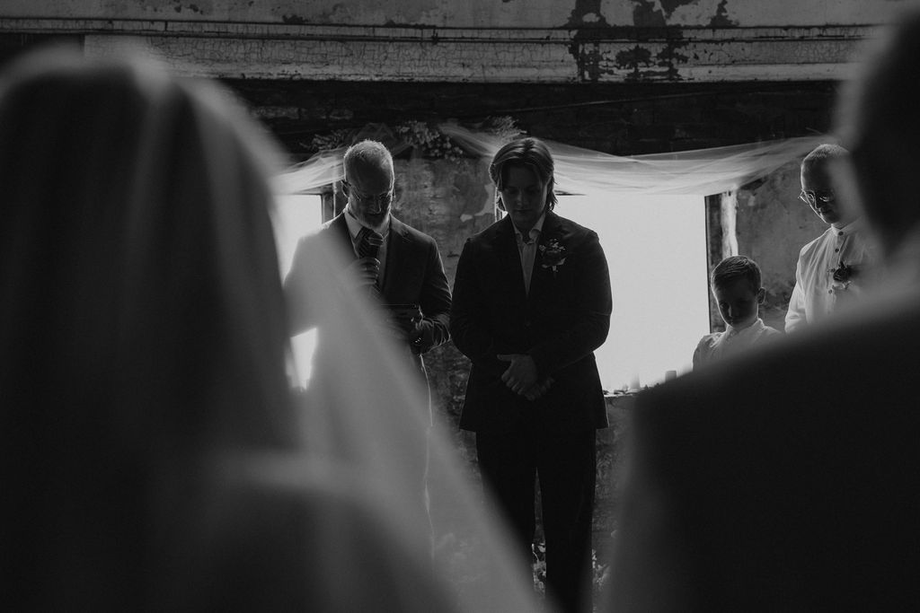 groom bows his head in prayer as his future bride stands in front of him at wedding ceremony at North Church venue in Muncie, Indiana.