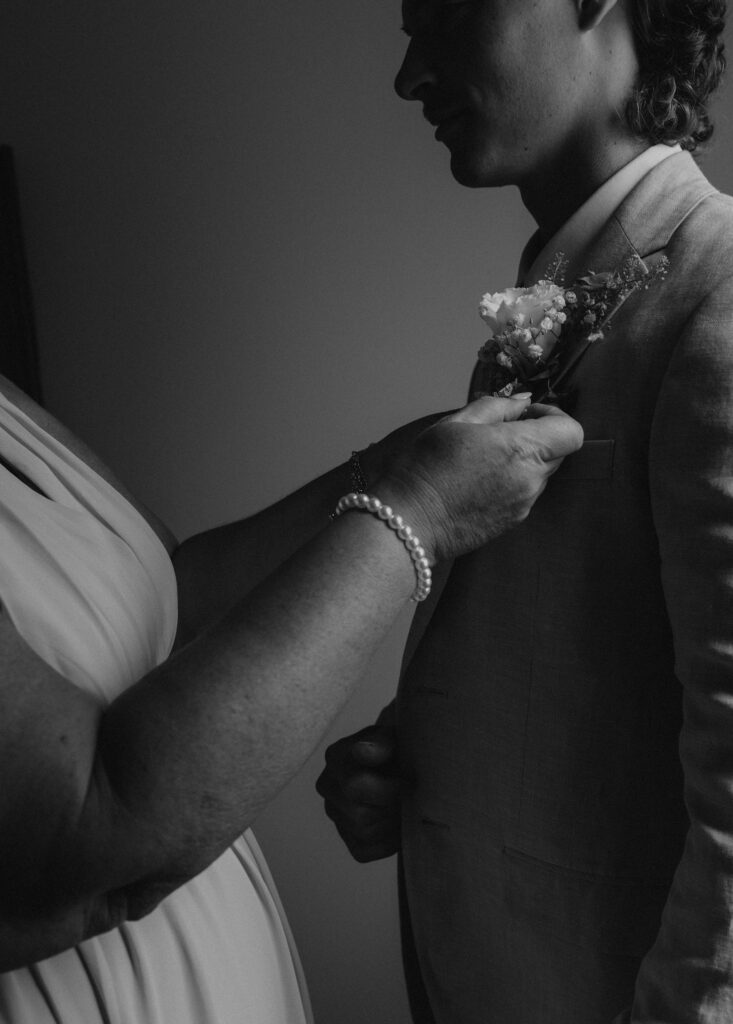 The groom in his outfit as he gets ready for the wedding day with the help of his mom, as she pins on his flowers.